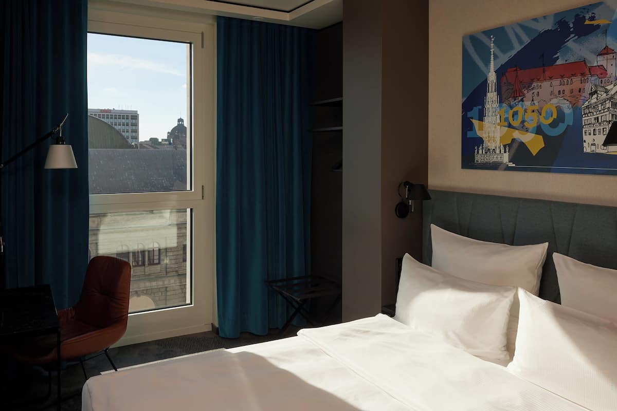 Een hotelkamer met een groot bed, een bureau met een lamp, een stoel en uitzicht op gebouwen door een raam.