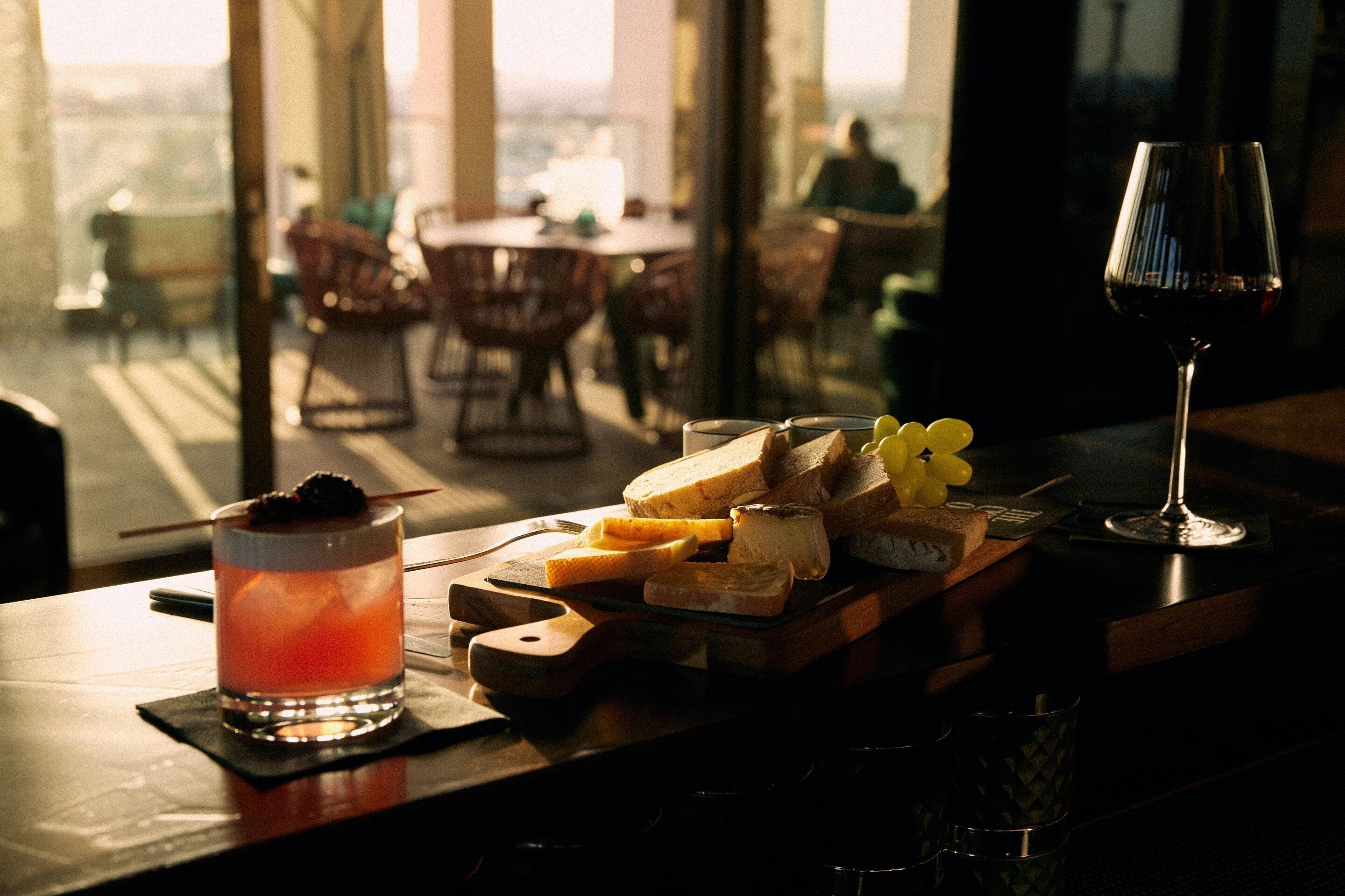 Eine Bar mit einem Weinglas Rotwein, einem Cocktail und verschiedenen Käsesorten auf einer Holzplatte.