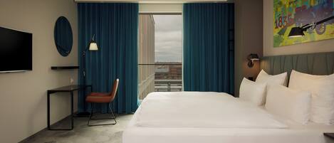 Una habitación de hotel moderna con una cama grande, un escritorio, una silla, un televisor y una ventana con cortinas.