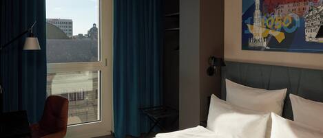 A hotel room with a large bed, a desk with a lamp, a chair, and a view of buildings through a window.