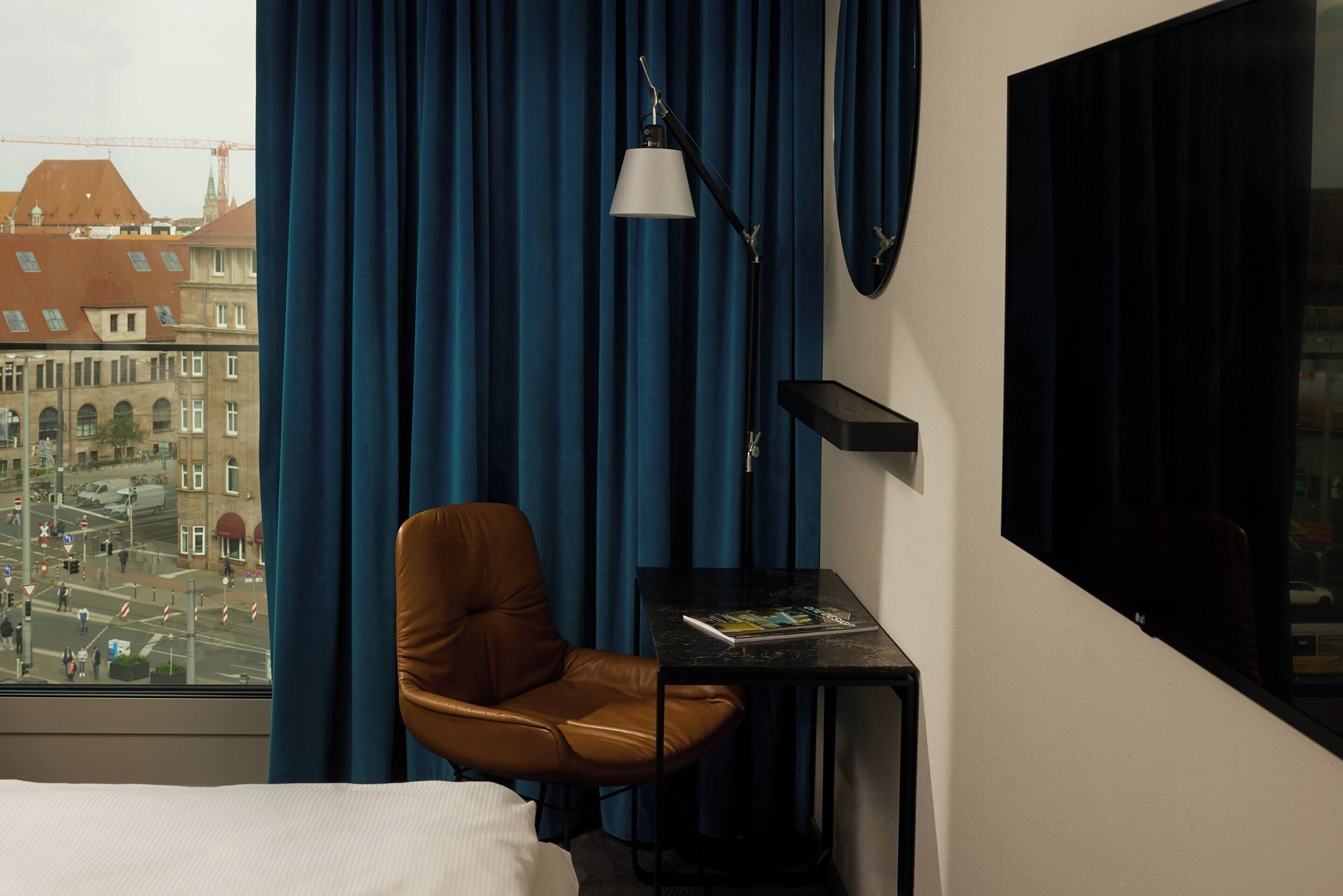 Habitación de hotel con vista a un paisaje urbano, un sillón de cuero marrón, una mesita con una revista y una lámpara negra.