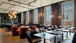 A modern restaurant with wooden tables and chairs, hanging pendant lights, and large windows offering a city view. - The Cloud One Nürnberg, by the Motel One Group (Nuremberg)