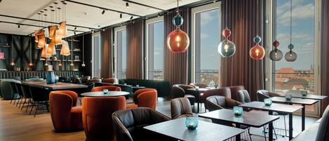 A modern restaurant with wooden tables and chairs, hanging pendant lights, and large windows offering a city view.