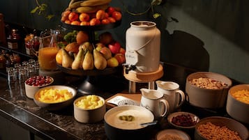 Una mesa de buffet con diversas opciones de comida, incluyendo frutas, un dispensador de bebidas y tazones de cereal y otros productos para el desayuno.