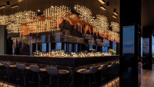 A bar area with a marble countertop, multiple stools, and a cityscape mural in the background. - The Cloud One Nürnberg, by the Motel One Group (Nuremberg)