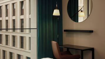 A hotel room with a bed, a desk, a chair, a mirror, a lamp, and a view of a building with windows.