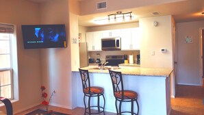 A modern kitchen with white cabinets, a microwave, and a built-in oven. There are two bar stools and a small island with a sink. A flat-screen TV is mounted on the wall. - Sun Ray #1A - Pet Friendly, 3min Walk to Beach (South Padre Island)