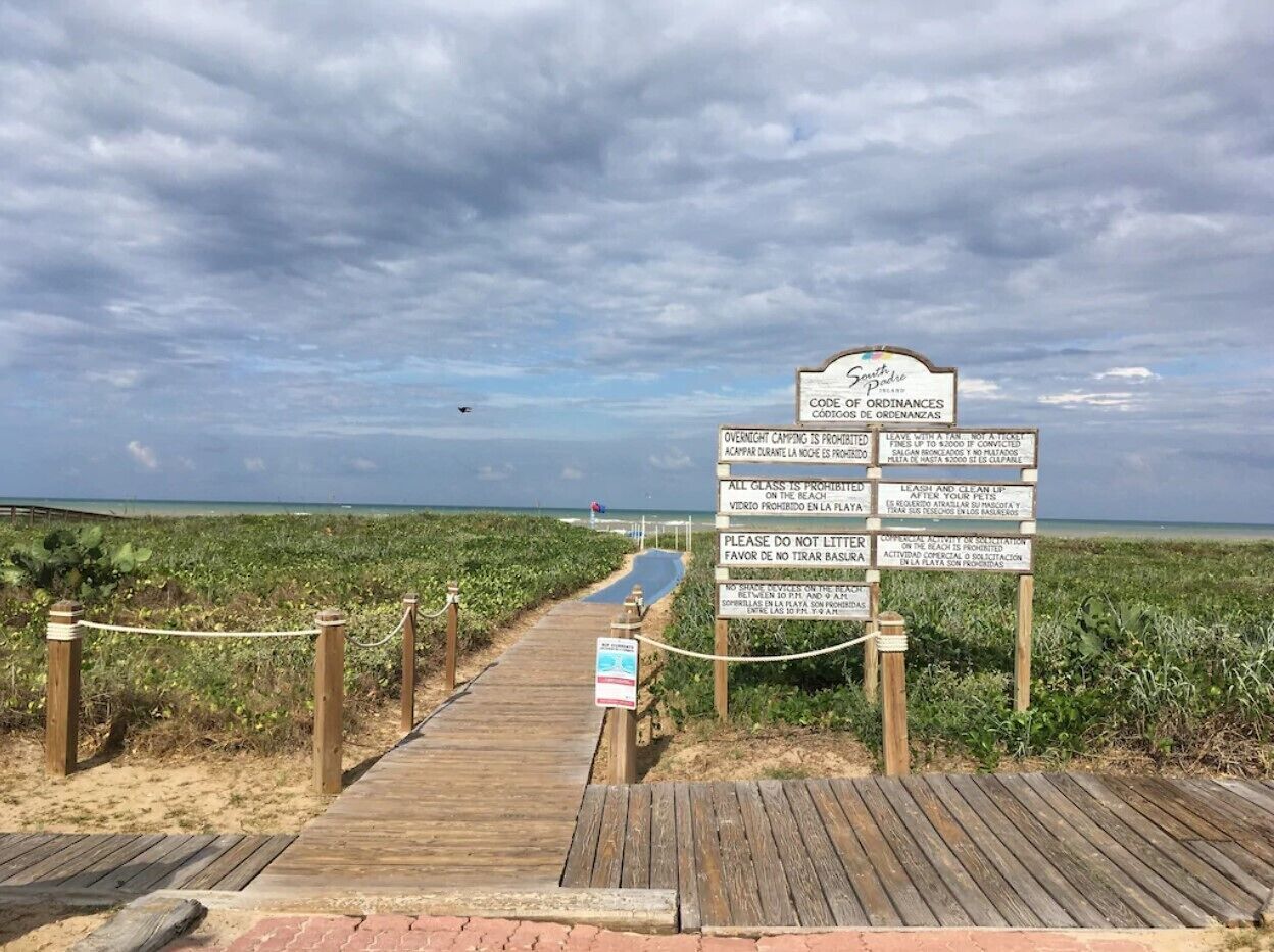 A wooden boardwalk leading to a beach area with a signboard displaying various beach rules and regulations.