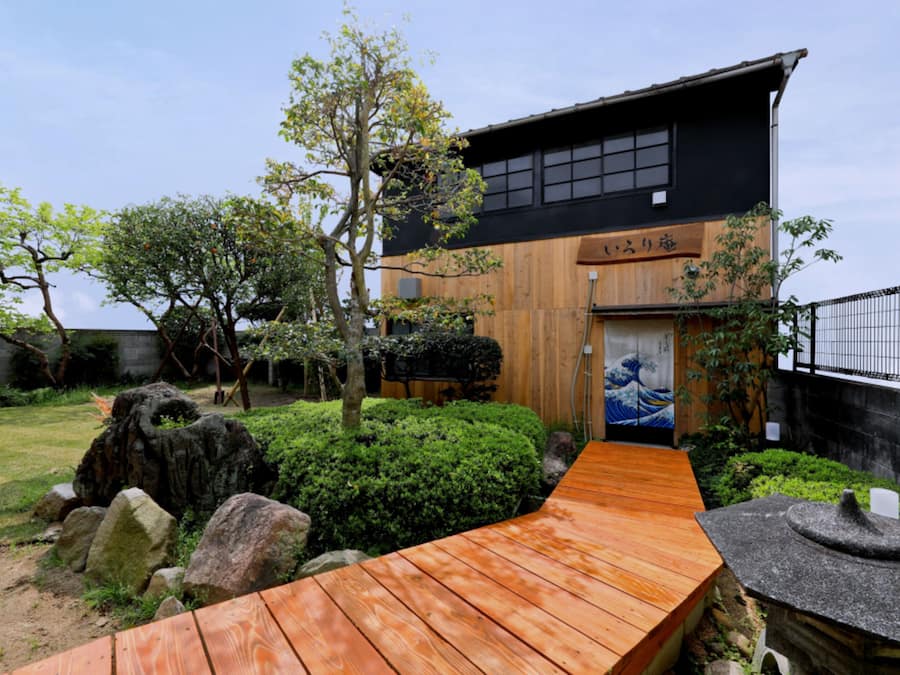 A traditional Japanese house with a wooden exterior, a stone pathway leading to the entrance, and a small garden with rocks and shrubs.