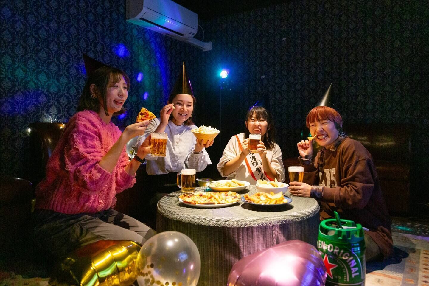 A group of people sitting around a table with food and drinks, in a room with patterned wallpaper and an air conditioner.