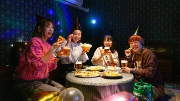A group of people sitting around a table with food and drinks, in a room with patterned wallpaper and an air conditioner.