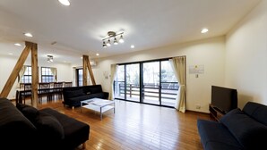 A spacious living room with a wooden floor, a black sofa, a coffee table, a TV, and a balcony with large windows.