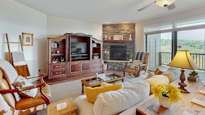 A spacious living room with a fireplace, comfortable seating, a television, and a large window offering a view. - Relaxing condo with sweeping valley views, deck, and fireplace (Sugar Mountain)
