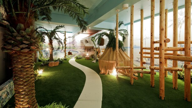 Un espacio interior con temática tropical que presenta un sendero serpenteante, palmeras, una hamaca colgante y una estructura de juego de madera.