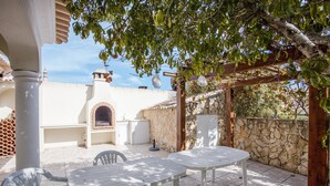 Un patio al aire libre con mesas y sillas blancas, un muro de piedra y una pérgola con luces colgantes.