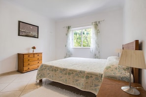 Une chambre à coucher avec un lit, une commode, une lampe, une fenêtre avec des rideaux et un tableau au mur.