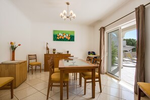 Un coin repas avec une table en bois, des chaises, un lustre, un tableau représentant des tulipes et une vue à travers une porte vitrée.
