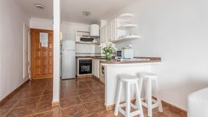 Una cocina con una isla central, taburetes blancos tipo bar, una puerta de madera y suelo de baldosas.
