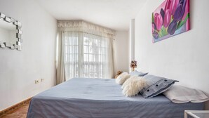 Un dormitorio con cama, espejo, un cuadro y ventana con cortinas.