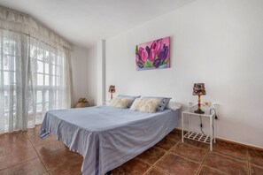 A bedroom with a bed, curtains, a painting, a lamp, and a small table.