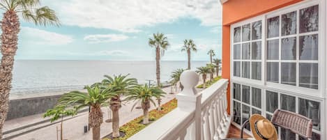 Un balcón con vista al mar, palmeras y una mesa de vidrio con un sombrero.