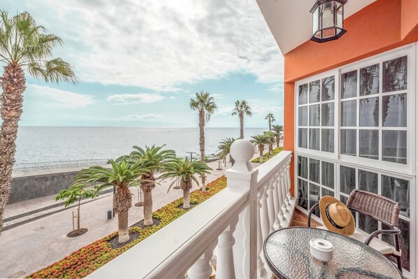 A balcony with a view of the sea, palm trees, and a glass table with a hat.