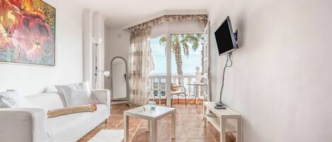 A living room with a white sofa, a small table, a TV mounted on the wall, and a view of the balcony.
