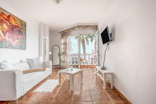 A living room with a white sofa, a small table, a TV mounted on the wall, and a view of the balcony.