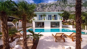 Una villa moderna con piscina, rodeada de palmeras y montañas al fondo.