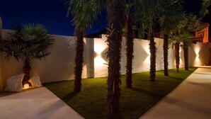 Un espacio exterior bien iluminado con palmeras, un muro blanco y un edificio de piedra al fondo.