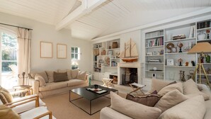 Uma sala de estar com sofá, poltronas, mesa de centro e uma estante repleta de livros e objetos decorativos.