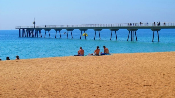 Une plage avec une jetée qui s’avance dans l’océan, des gens assis sur le sable et un ciel bleu dégagé.