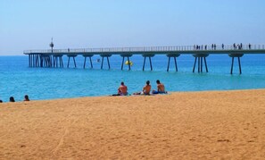 A beach with a pier extending into the ocean, people sitting on the sand, and a clear blue sky. - Casa Luna, junto playa y Barcelona Centro. (Badalona)