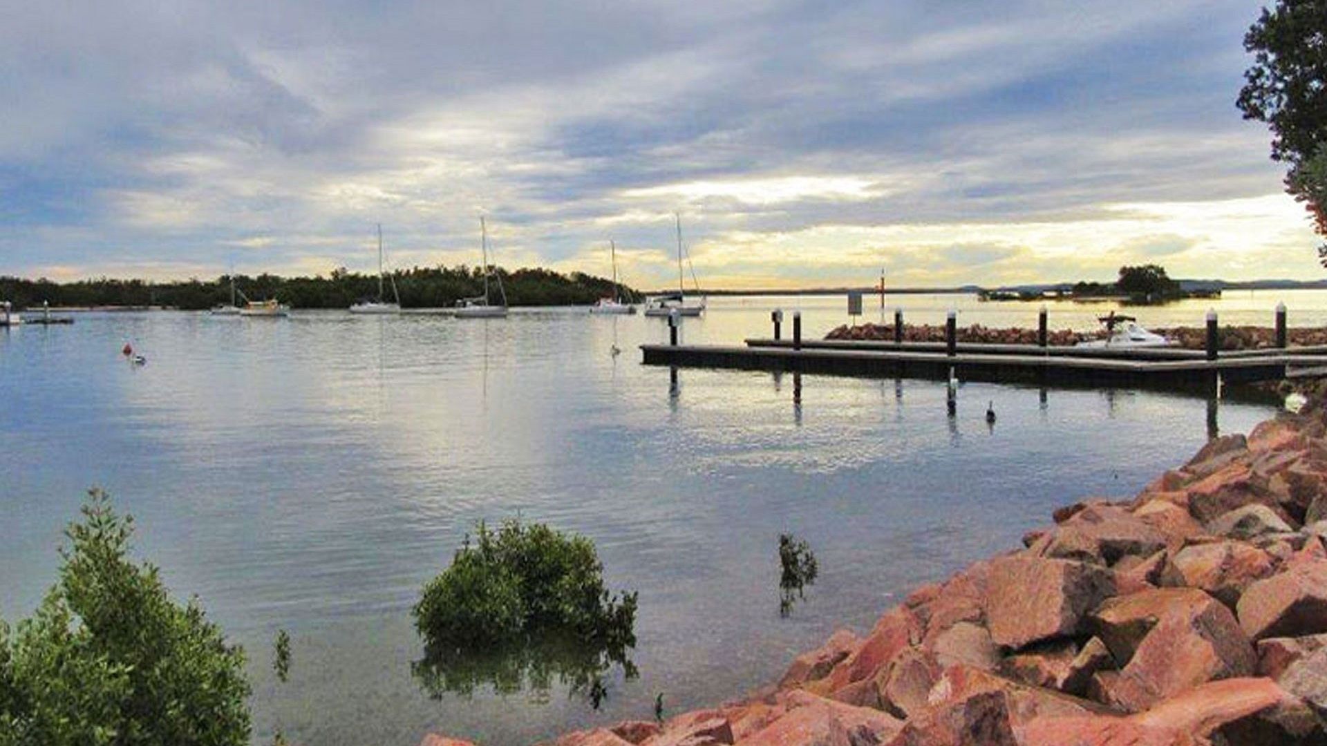 A calm waterfront with boats docked, a pier extending into the water, and a rocky shoreline with greenery.