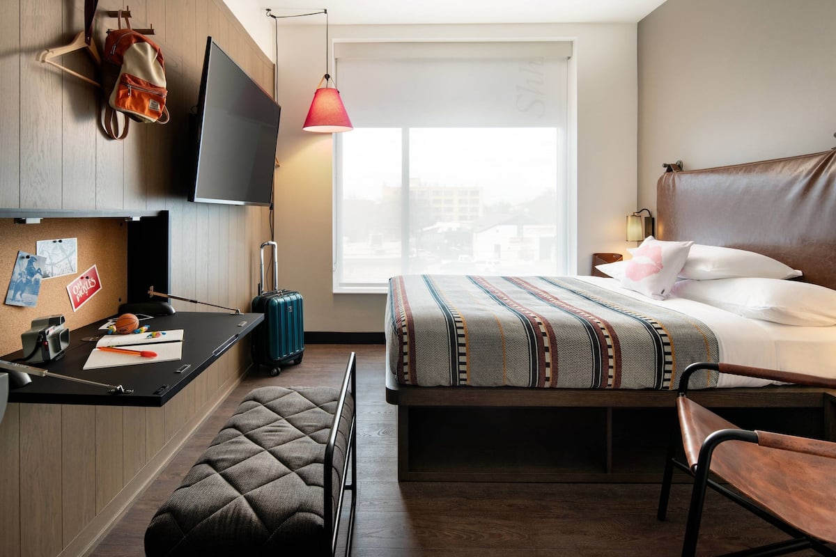 A hotel room with a bed, a desk with a computer, a chair, a suitcase, and a TV.