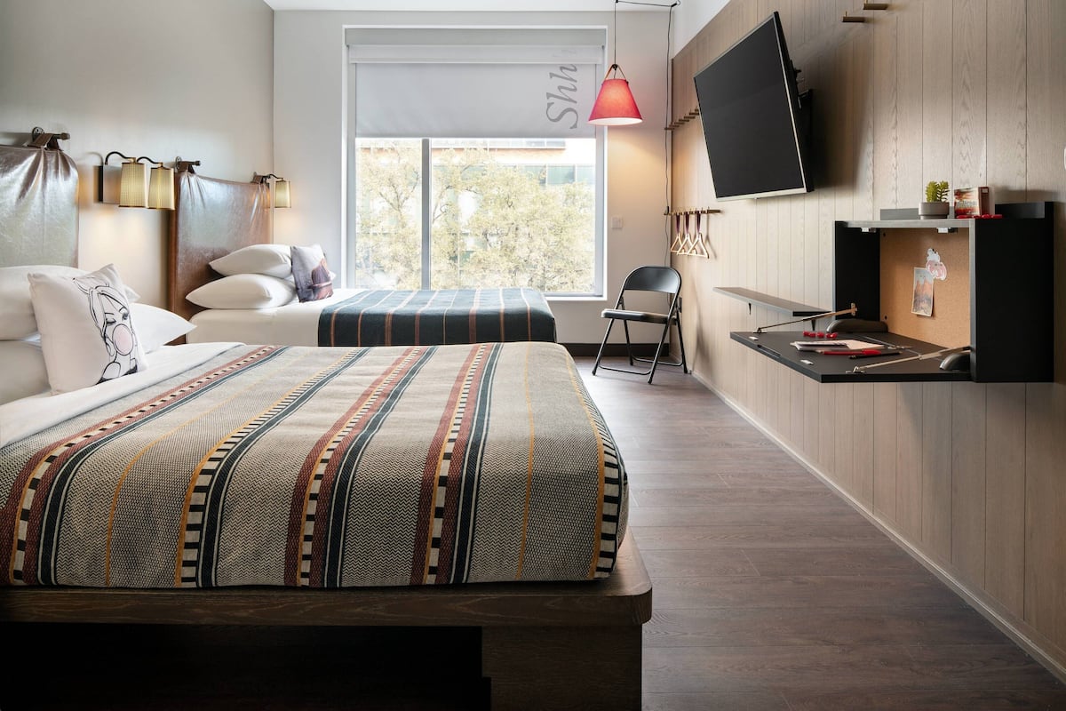 A modern hotel room with two beds, a TV, a desk, and a window with a view of trees.
