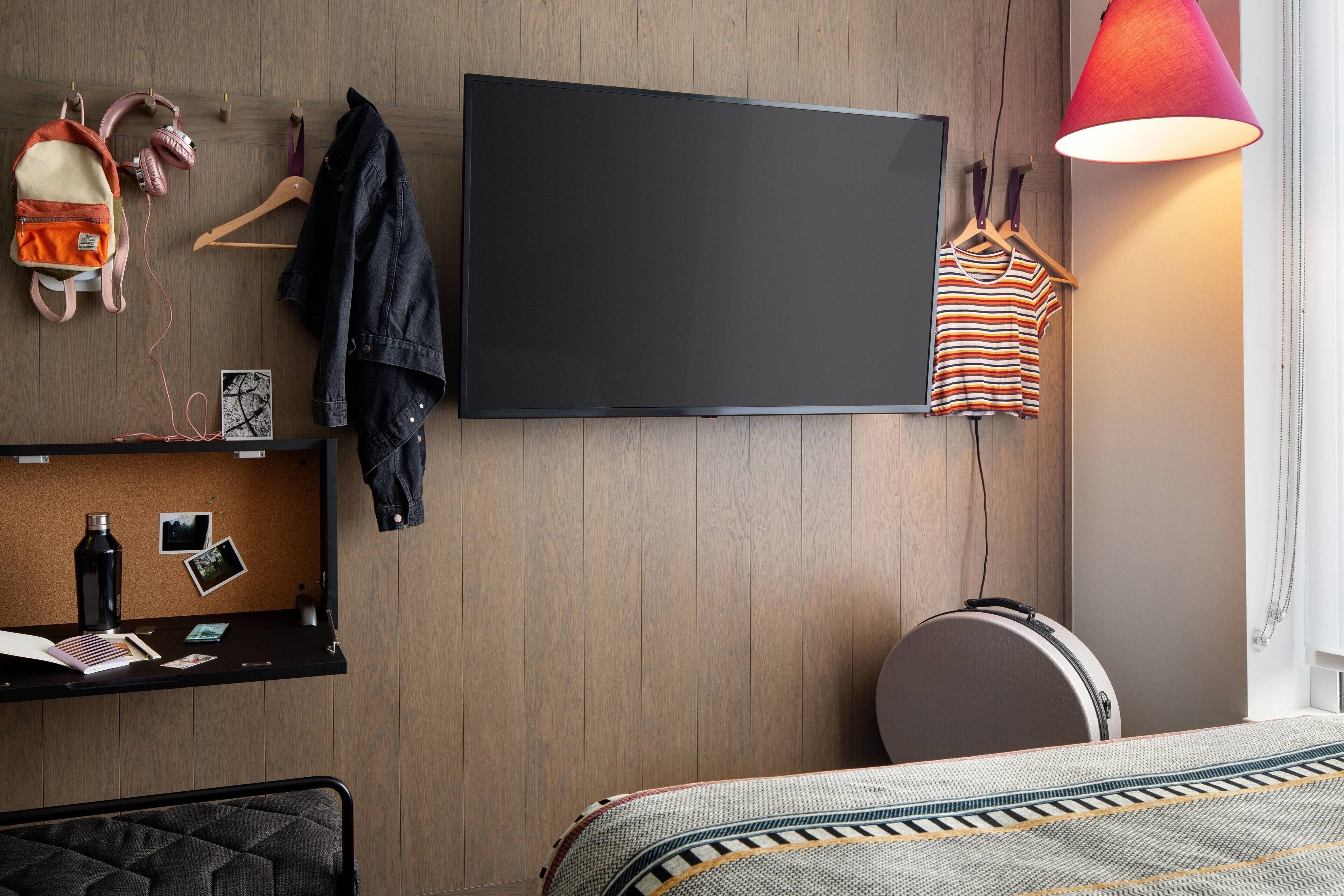 Ein modernes Hotelzimmer mit einem großen Flachbildfernseher, einer Holzvertäfelung, einem Schreibtisch mit einem Laptop, einem Koffer und einem Bett mit einem gemusterten Überwurf.