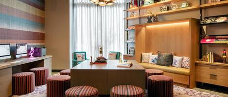 A modern, well-lit room with a central table surrounded by striped ottomans, a bookshelf with decorative items, and a large window with sheer curtains.
