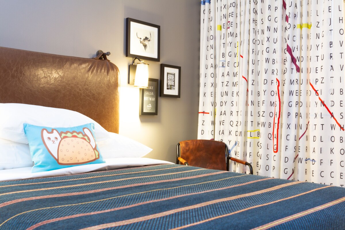 A hotel room with a large bed, a headboard, a pillow with a cat design, a framed picture, a lamp, and a chair.