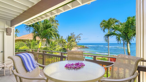 A balcony with a round table and two chairs, overlooking a beach with palm trees and ocean views. - Sunrise Serenity| 2BR Oceanfront Condo With AC (Kapaa)