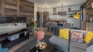 Ein modernes Wohnzimmer mit Flachbildfernseher, einer Couch mit dekorativen Kissen, einem Couchtisch mit Flasche und Kerzen sowie einem Essbereich mit Tisch und Stühlen.