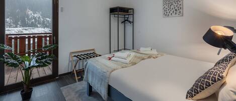 Un dormitorio con cama, lámpara de noche, lámpara colgante, ventana con vista a árboles cubiertos de nieve, una planta y un perchero.