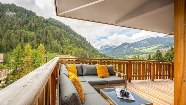 Ein Balkon mit Holzdeck, ein graues Sofa mit gelben Kissen und ein Tisch mit zwei Tassen und einem Löffel.