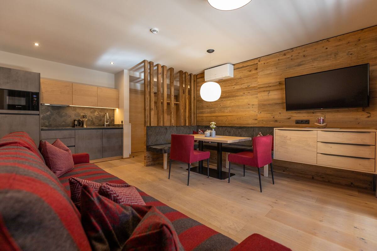A modern kitchen with wooden accents, a dining area with red chairs, and a large flat-screen TV mounted on the wall.