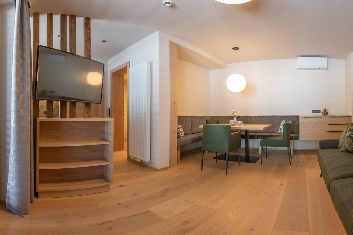 A modern living room with a wooden floor, a green sofa with patterned cushions, a dining area with a table and chairs, and a TV mounted on the wall.