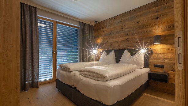 A bedroom with a wooden headboard, a bed with white linens, a bedside table, and a window with blinds. - Sport Apart (Zell am Ziller)