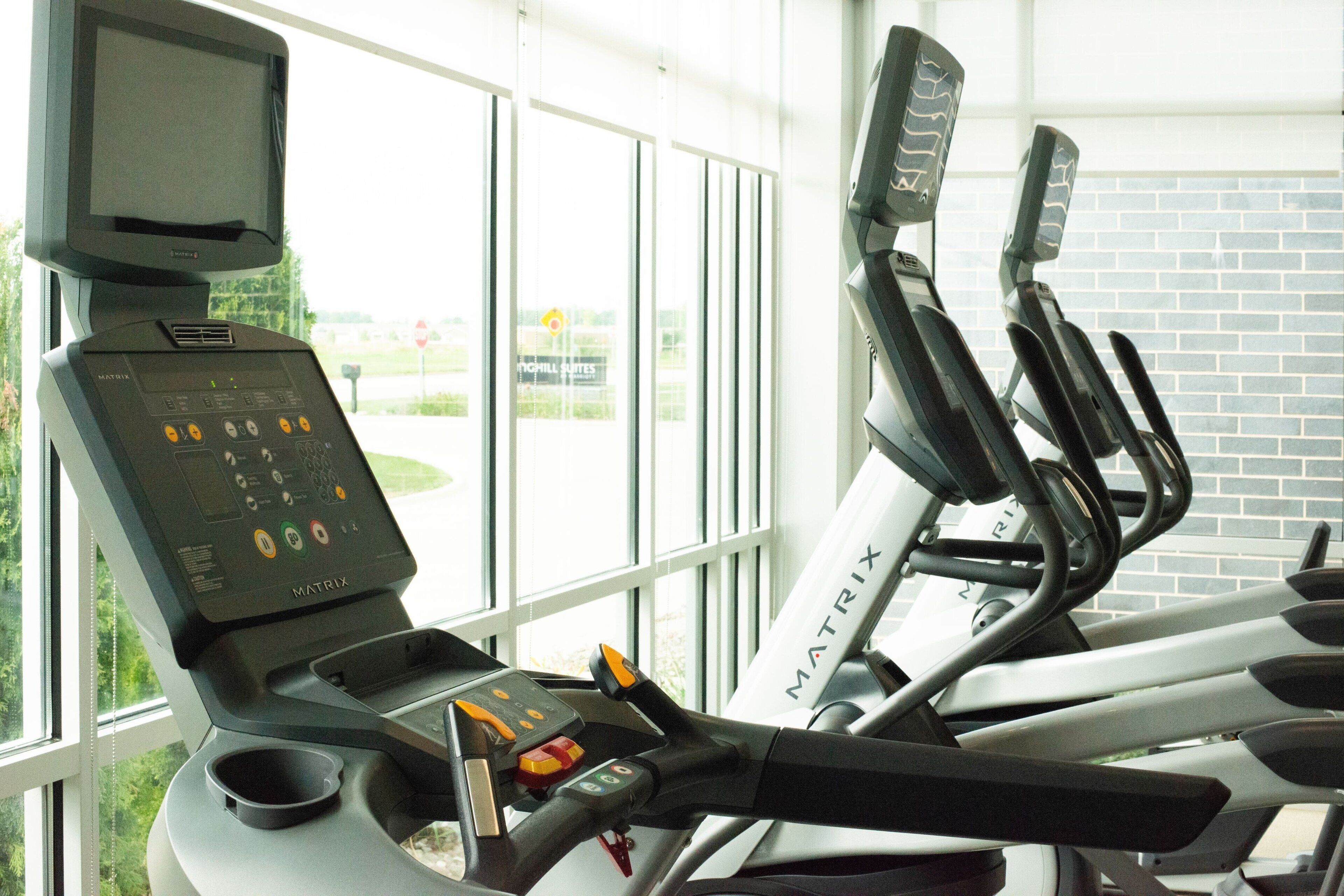 A treadmill with a digital display and control panel, positioned near a window with a view of a brick wall and greenery outside.