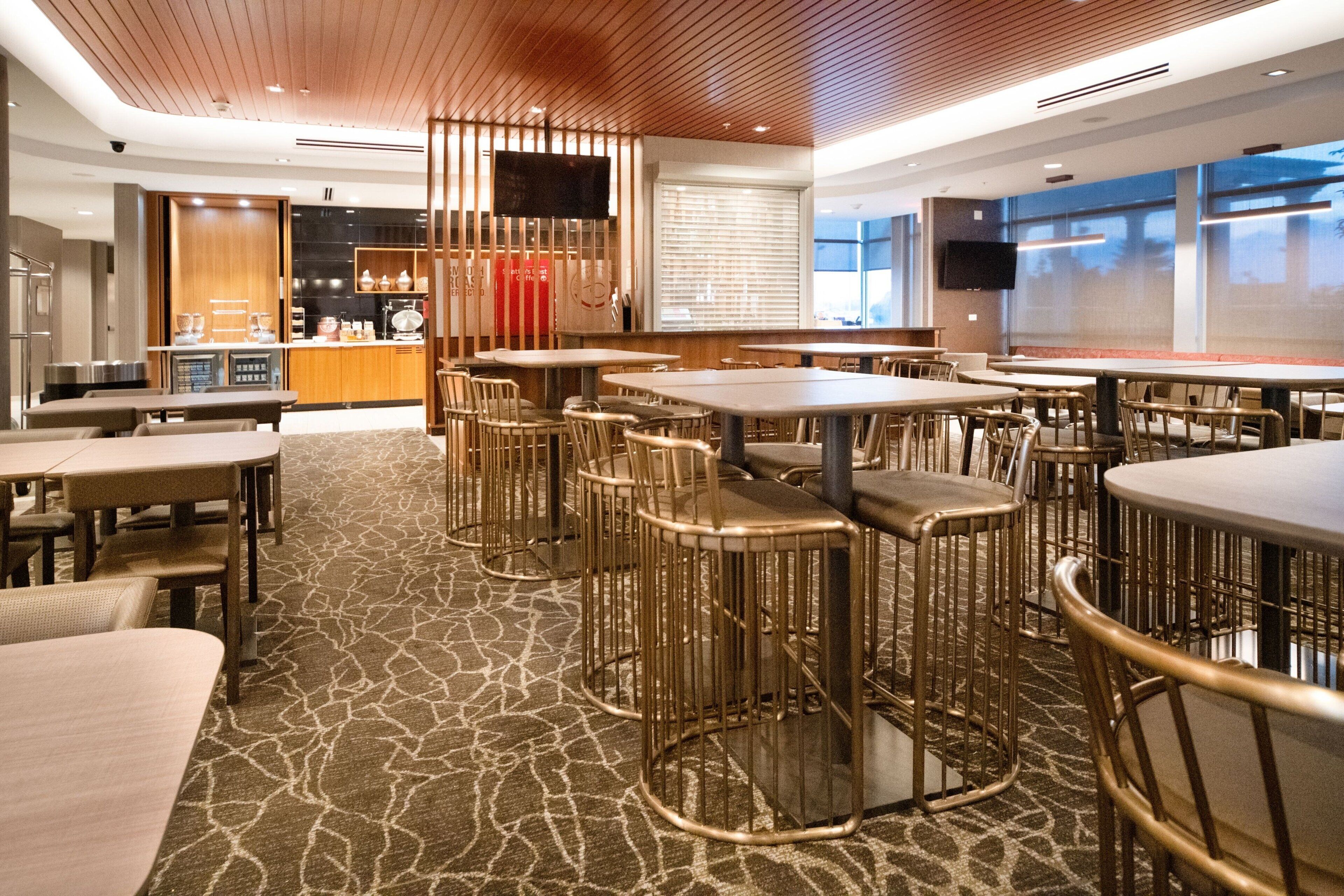 Un restaurante moderno con mesas altas y taburetes, una zona de cocina y un amplio ventanal con persianas.