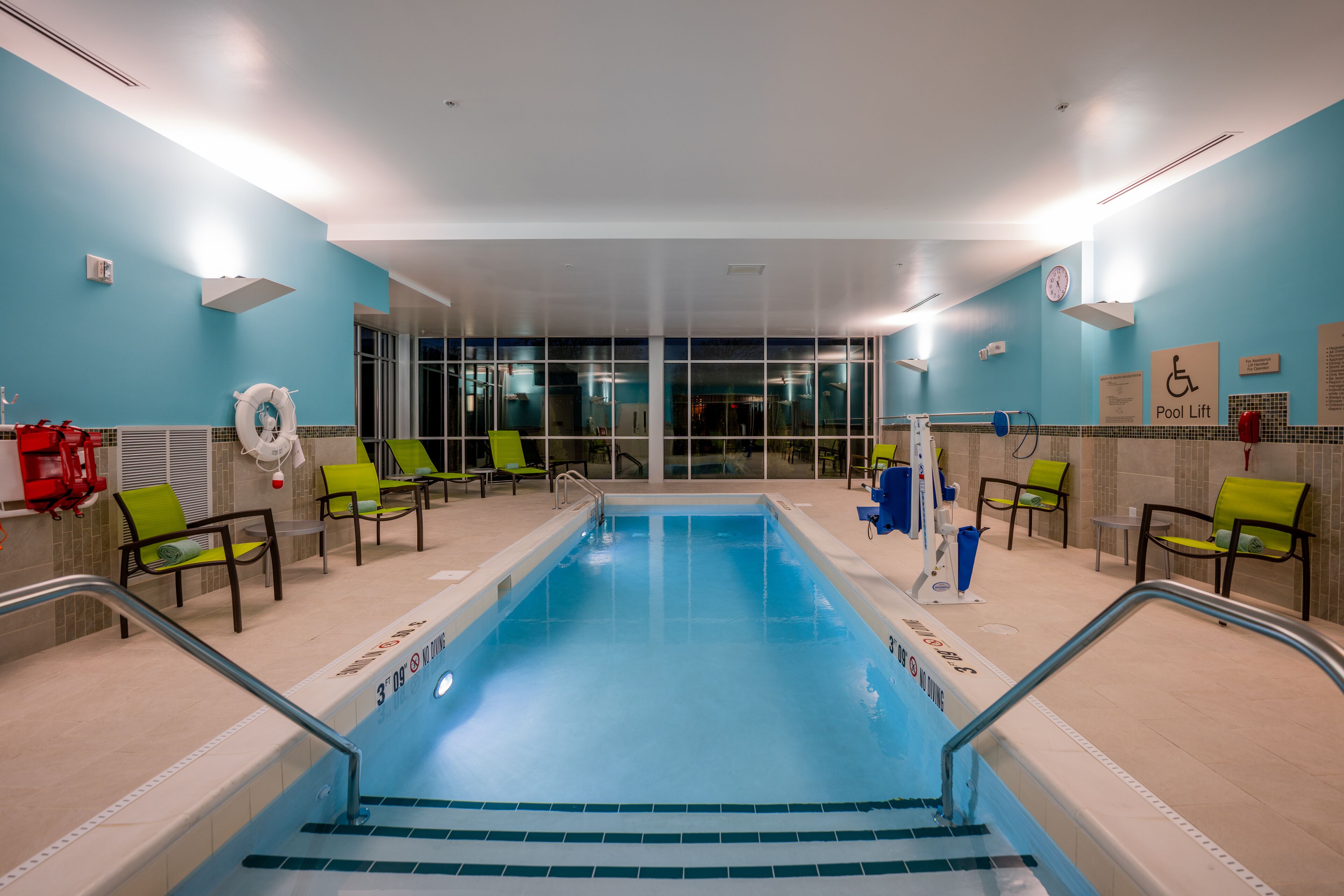 Una piscina con suelo de baldosas azules, rodeada de sillones de descanso verdes y negros, y un cartel de elevador para piscina en la pared.