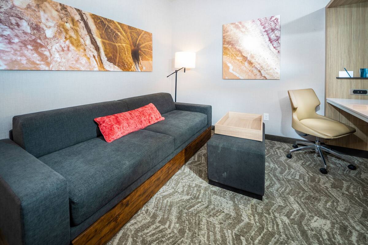 A modern hotel room with a grey sofa, a red pillow, a wooden side table, a beige chair, and a desk with a lamp.